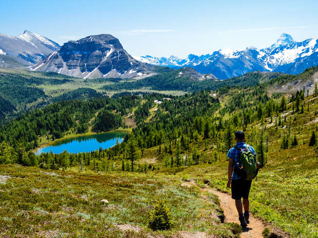 A hiker with a backpack walks along a narrow trail overlooking a turquoise lake, surrounded by lush greenery and distant snow-capped mountains under a clear blue sky.
